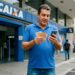 Trabalhador confere o celular e segura cédulas em frente à Caixa, em referência ao Abono Salarial de junho de 2026
