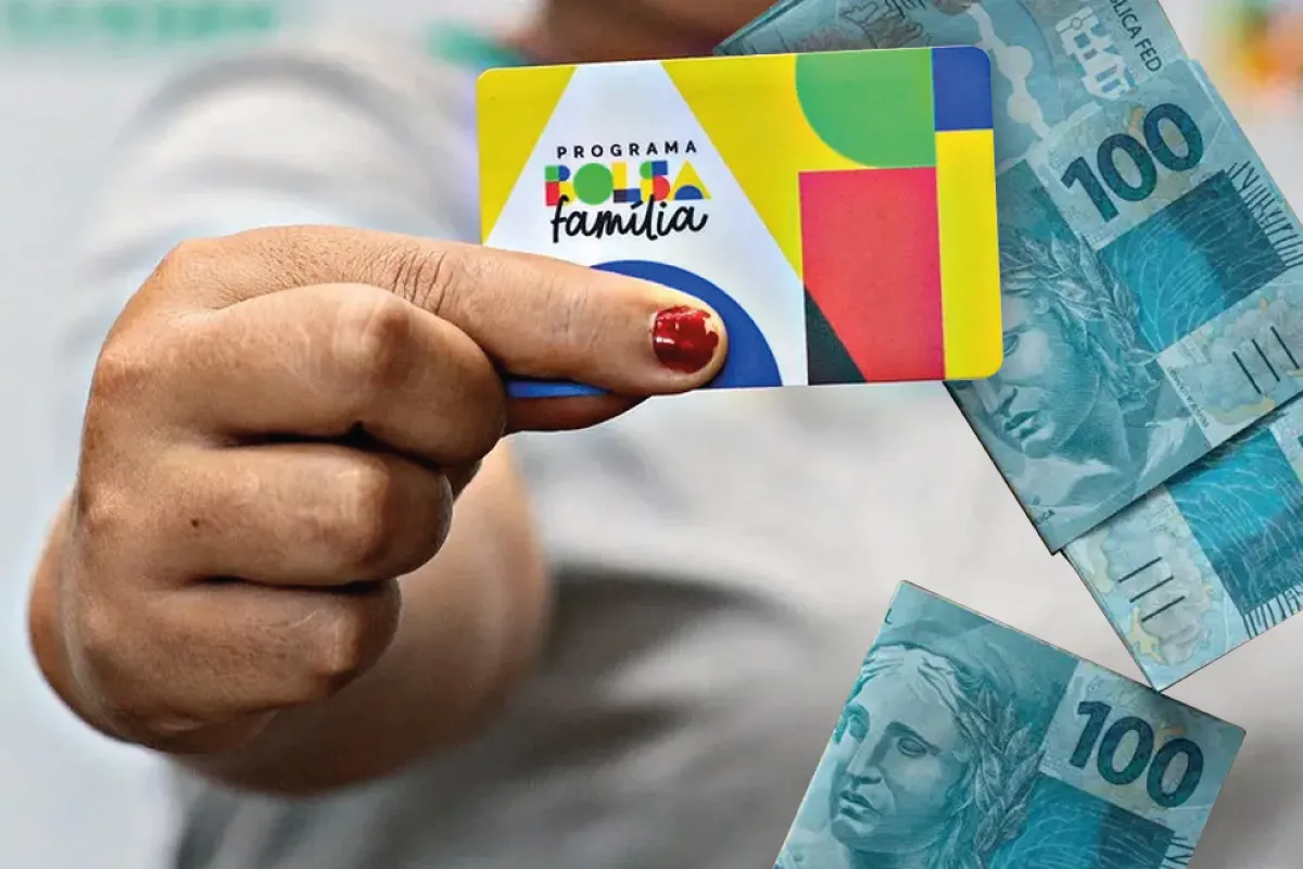 Mão segura o cartão do Programa Bolsa Família ao lado de cédulas de R$ 100, ilustrando o pagamento antecipado de junho de 2026