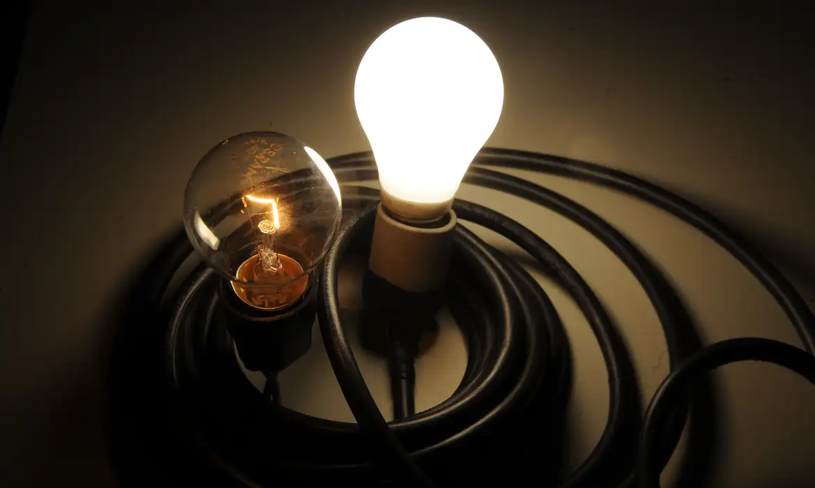 Duas lâmpadas acesas lado a lado, uma incandescente e outra LED, sobre fios enrolados