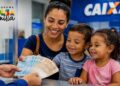 Imagem ilustrativa representa mãe sorridente com dois filhos pequenos recebendo notas de dinheiro de um atendente no balcão de uma agência da Caixa, ao lado do logo do Programa Bolsa Família