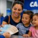 Imagem ilustrativa representa mãe sorridente com dois filhos pequenos recebendo notas de dinheiro de um atendente no balcão de uma agência da Caixa, ao lado do logo do Programa Bolsa Família