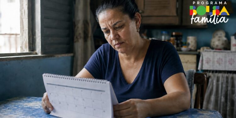 Mulher sentada à mesa consulta um calendário, ao lado da logomarca do Programa Bolsa Família, em referência às datas de pagamento de junho de 2026