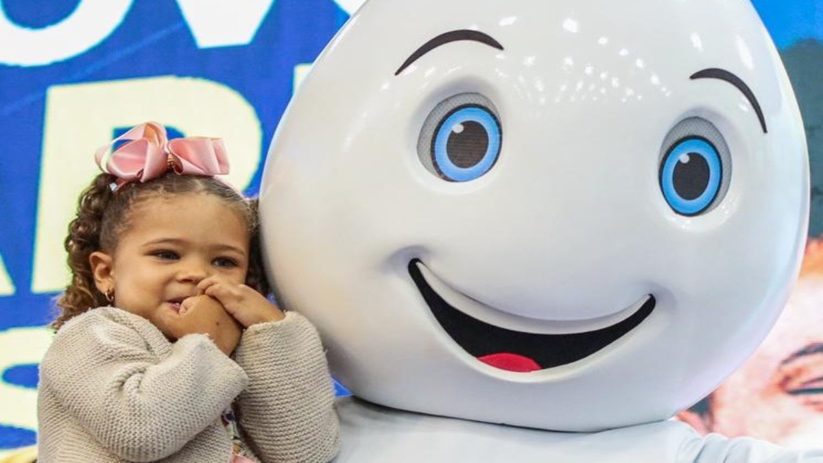 Criança sorridente abraçada a um mascote em ação de saúde, representando o impacto do Bolsa Família na saúde dos beneficiários.