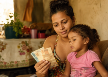 Mãe segura notas de cem e cinquenta reais ao lado da filha pequena sentada no colo