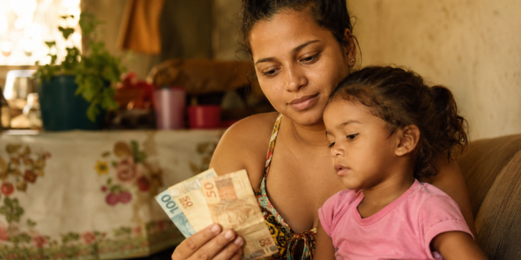 Mãe segura notas de cem e cinquenta reais ao lado da filha pequena sentada no colo