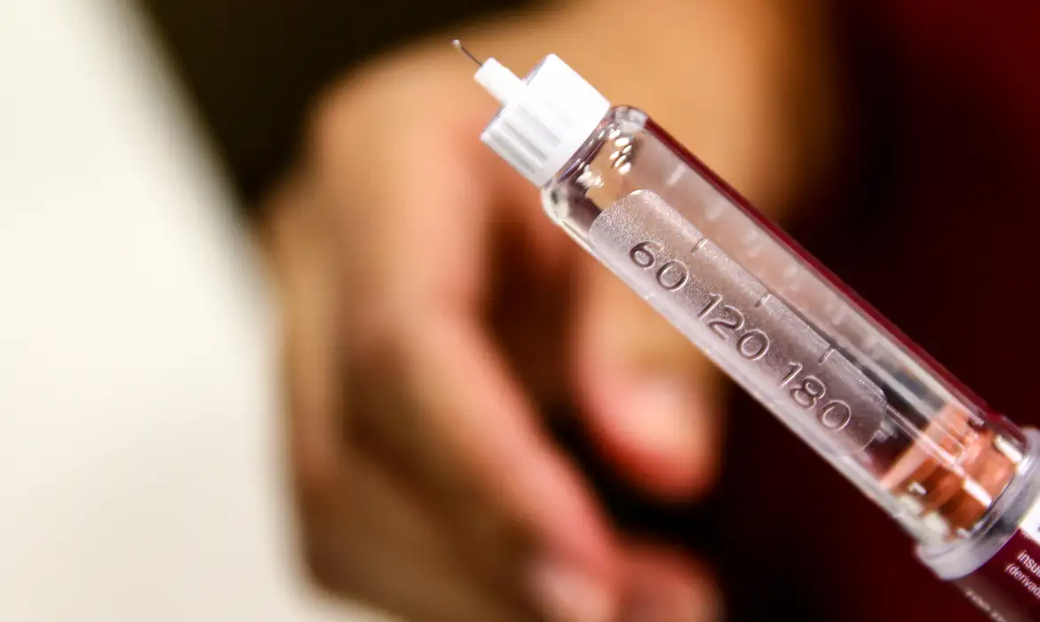 lose de uma caneta injetável transparente com escala de dosagem, segurada perto da mão de uma pessoa, em referência à autoaplicação do medicamento no projeto do SUS