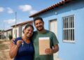 Casal sorri segurando chave e envelope em frente a casa popular azul recém-recebida