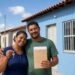 Casal sorri segurando chave e envelope em frente a casa popular azul recém-recebida