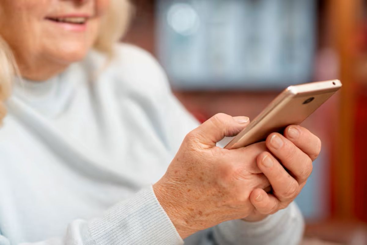 Mulher idosa segura celular com as duas mãos enquanto sorri em ambiente interno