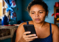 Beneficiária consulta o celular com atenção dentro de casa