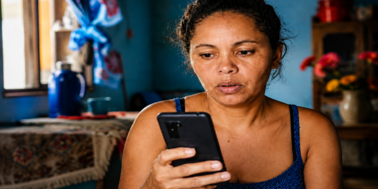 Beneficiária consulta o celular com atenção dentro de casa