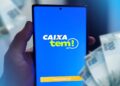 Mão segura celular com o aplicativo Caixa Tem aberto, com cédulas de real ao fundo.