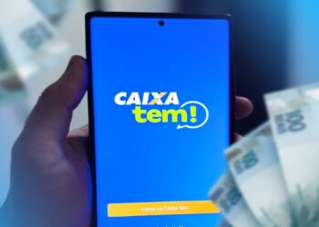 Mão segura celular com o aplicativo Caixa Tem aberto, com cédulas de real ao fundo.