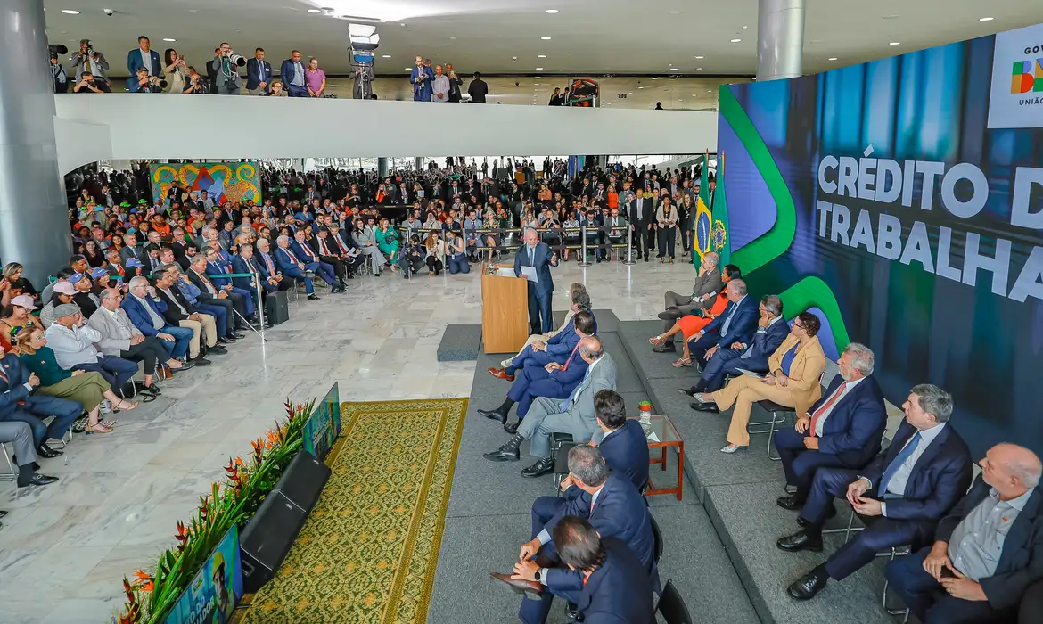Cerimônia do governo federal sobre o Crédito do Trabalhador, com autoridade no púlpito, plateia e painel do programa ao fundo