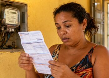 Mulher confere a conta de luz ao lado do medidor de energia, ilustrando o desconto do bônus de Itaipu na fatura de agosto
