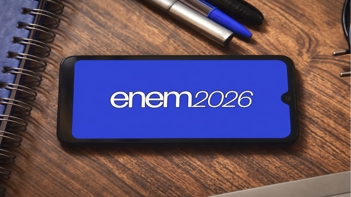 Celular sobre uma mesa de madeira exibindo a logo do Enem 2026, ao lado de canetas e caderno, representando a inscrição e o pagamento da taxa do exame.