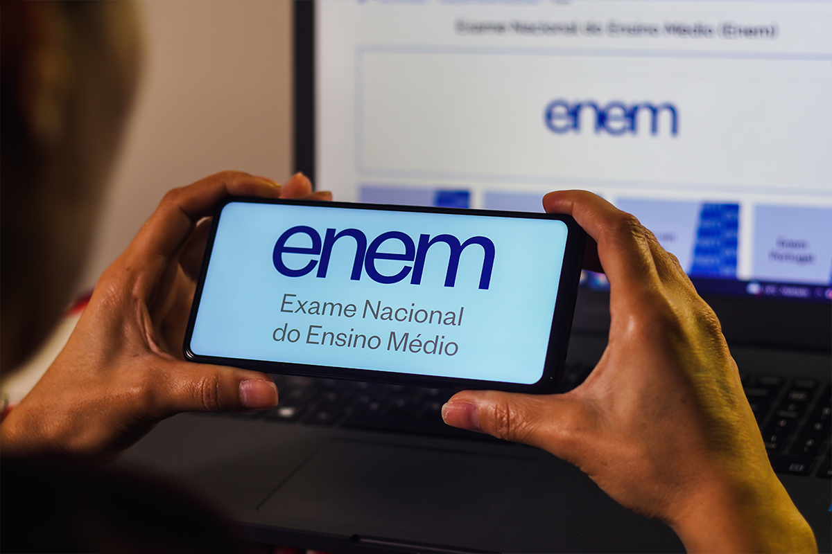 Pessoa segura celular com a logomarca do Enem diante do computador, em alusão à inscrição na Página do Participante