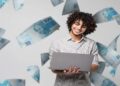 Jovem sorridente usa um notebook cercado por cédulas de R$ 100, representando as formas de pagamento da taxa de inscrição do Enem 2026.
