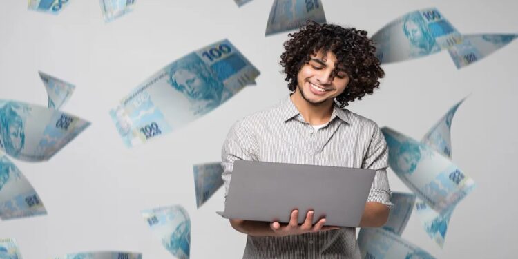Jovem sorridente usa um notebook cercado por cédulas de R$ 100, representando as formas de pagamento da taxa de inscrição do Enem 2026.