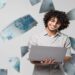 Jovem sorridente usa um notebook cercado por cédulas de R$ 100, representando as formas de pagamento da taxa de inscrição do Enem 2026.