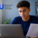 Jovem estudante de camiseta azul faz a inscrição no Sisu+ 2026 pelo notebook, segurando um documento, ao lado do logo do Sistema de Seleção Unificada