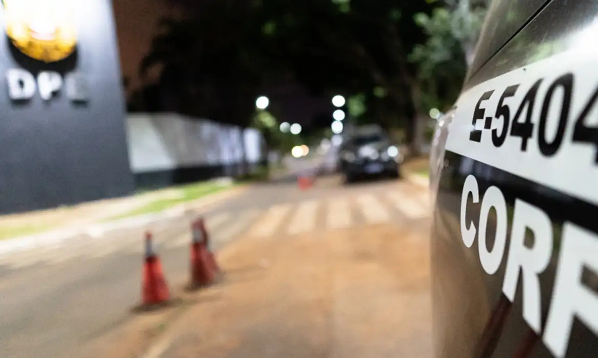 Viatura policial estacionada à noite diante de um prédio público, com cones de sinalização, durante a operação contra o PCC.