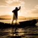 Pescador lança rede de pesca em barco ao pôr do sol em rio brasileiro
