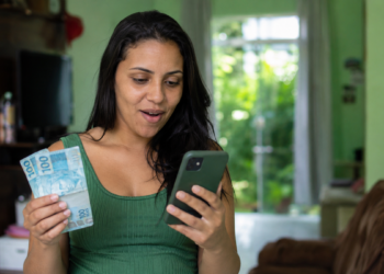 Mulher surpresa confere o celular segurando notas de cem reais
