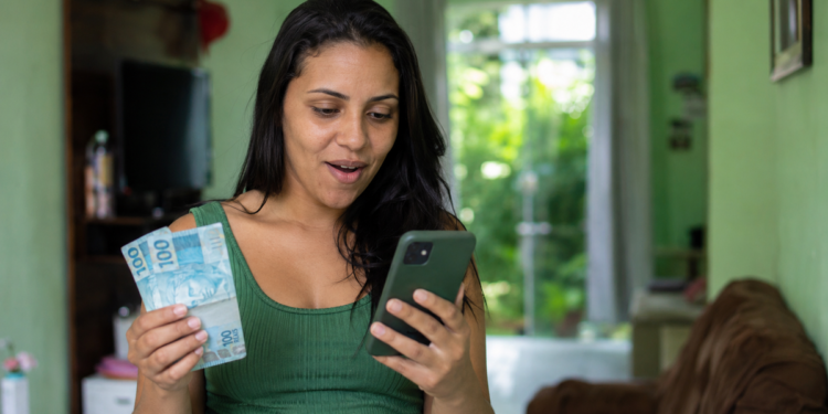 Mulher surpresa confere o celular segurando notas de cem reais