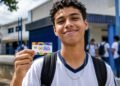 Estudante do ensino médio sorrindo e segurando o cartão do Pé-de-Meia em frente à escola, representando o pagamento do incentivo-frequência
