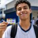 Estudante do ensino médio sorrindo e segurando o cartão do Pé-de-Meia em frente à escola, representando o pagamento do incentivo-frequência