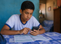Estudante de uniforme consulta o celular com documento sobre a mesa em casa