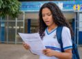 Estudante de uniforme e mochila lê um documento em frente à escola com expressão de dúvida, ao lado da logomarca do Pé-de-Meia