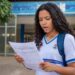 Estudante de uniforme e mochila lê um documento em frente à escola com expressão de dúvida, ao lado da logomarca do Pé-de-Meia