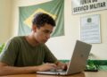 Jovem de camiseta verde faz o alistamento militar pelo notebook em uma sala da Junta de Serviço Militar, com a bandeira do Brasil ao fundo