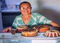 Confeiteira sorrindo atrás de um balcão com bolos e doces no próprio negócio, em referência aos empreendedores de baixa renda atendidos pelo Programa Acredita no Primeiro Passo