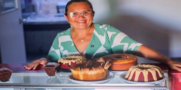 Confeiteira sorrindo atrás de um balcão com bolos e doces no próprio negócio, em referência aos empreendedores de baixa renda atendidos pelo Programa Acredita no Primeiro Passo