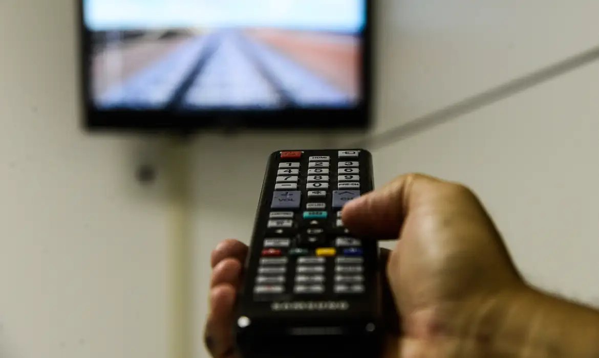 Mão aponta controle remoto para uma TV ligada, ilustrando o acesso à TV aberta pelo programa Brasil Antenado.