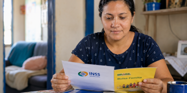 Mulher analisa documento do INSS e cartilha do Bolsa Família à mesa em casa