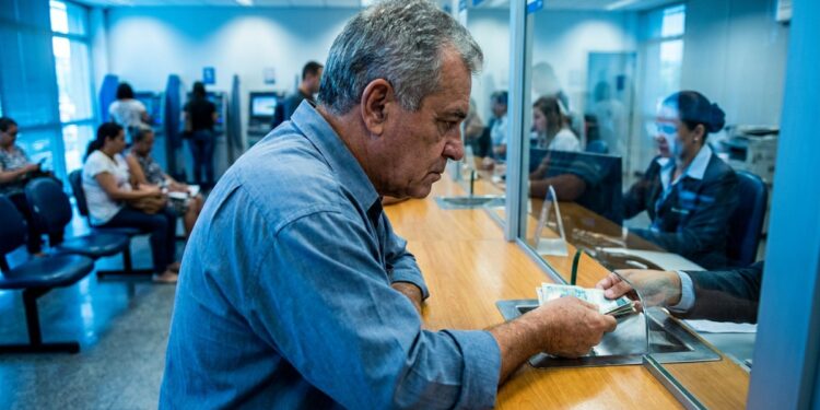 Aposentado confere cédulas de dinheiro no caixa de uma agência, representando a devolução dos descontos indevidos do INSS em 2026.