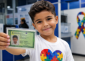 Menino de cabelo castanho segura carteira de identidade com símbolo do autismo, vestindo camiseta branca com quebra-cabeça colorido
