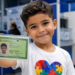 Menino de cabelo castanho segura carteira de identidade com símbolo do autismo, vestindo camiseta branca com quebra-cabeça colorido