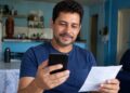 Homem sorrindo consulta o celular segurando um comprovante em casa, verificando a restituição do Imposto de Renda