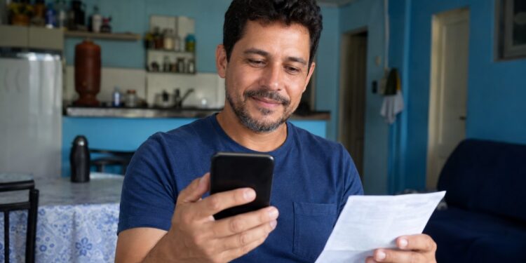 Homem sorrindo consulta o celular segurando um comprovante em casa, verificando a restituição do Imposto de Renda