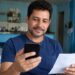 Homem sorrindo consulta o celular segurando um comprovante em casa, verificando a restituição do Imposto de Renda
