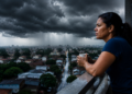 Mulher observa nuvens carregadas na varanda com vista para cidade durante a chuva