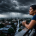 Mulher observa nuvens carregadas na varanda com vista para cidade durante a chuva