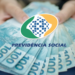 Mãos segurando notas de 100 reais em leque sob logo da Previdência Social do Brasil.
