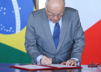 Autoridade masculina de aproximadamente 60-70 anos, vestindo terno cinza e gravata azul estampada, assina documento oficial à mesa.