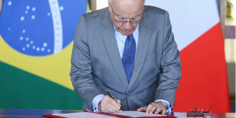 Autoridade masculina de aproximadamente 60-70 anos, vestindo terno cinza e gravata azul estampada, assina documento oficial à mesa.